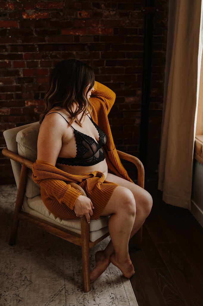 A woman sits on a chair looking out the window with her hands sitting on her lap.  She is wearing a matching lace set of lingerie and a chunky knit orange sweater.