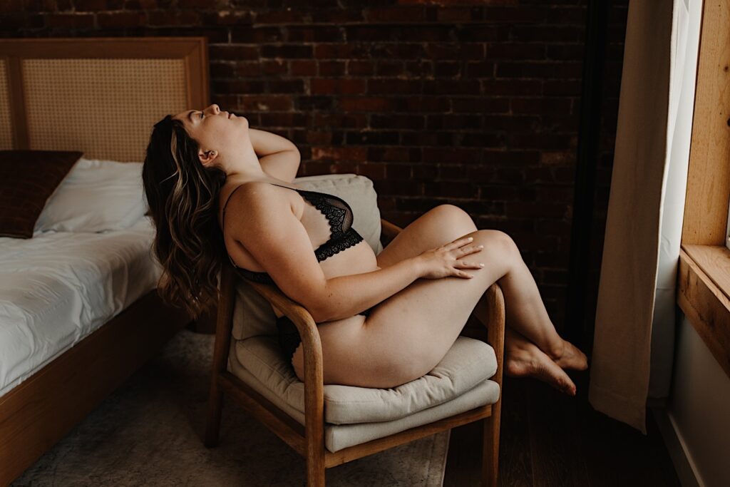 A woman sits with her head back and her legs draped over the edge of the chair in a black two piece set of lingerie.