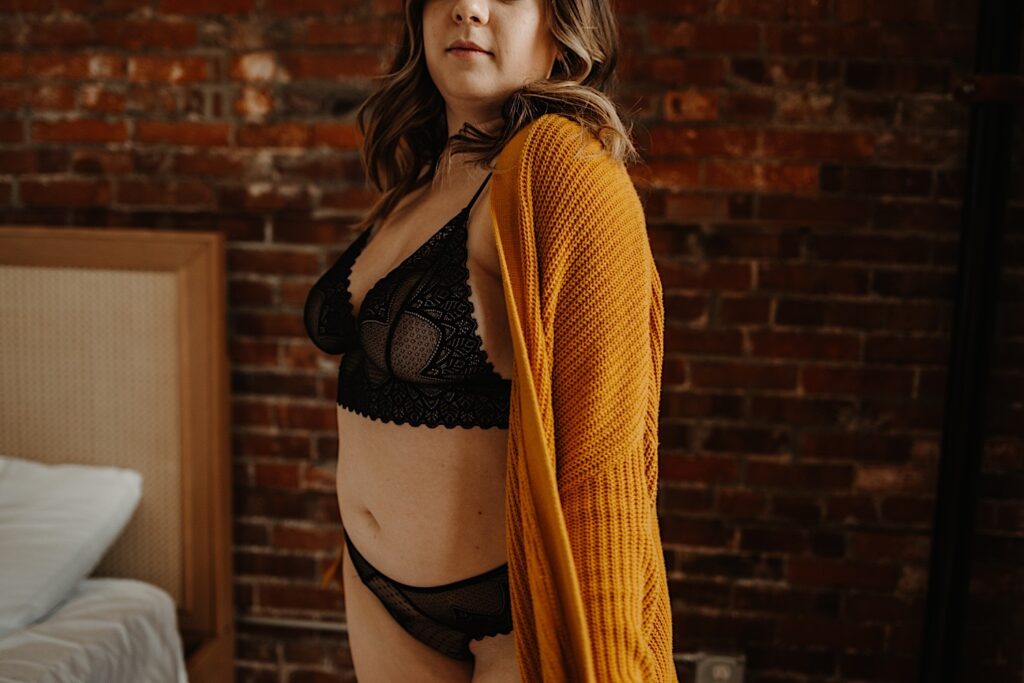 A woman stands facing the camera wearing a two piece set of lingerie and wearing an orange chunky knit sweater.