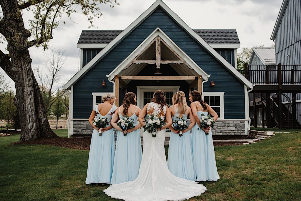 A bride and bridesmaids stand away from the camera holding their bouquets behind their backs in front of the getting ready house at 1841 Farms and Vineyard