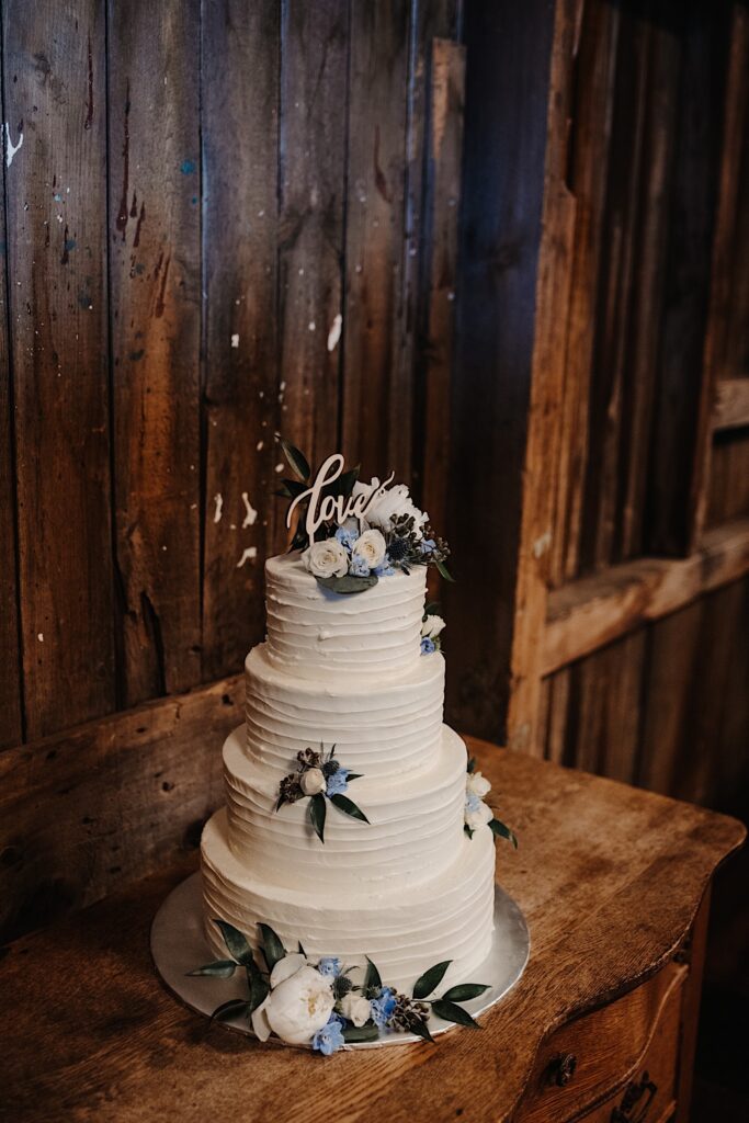 A wedding cake on a wooden table with white and blue flowers as decorations throughout the cake.