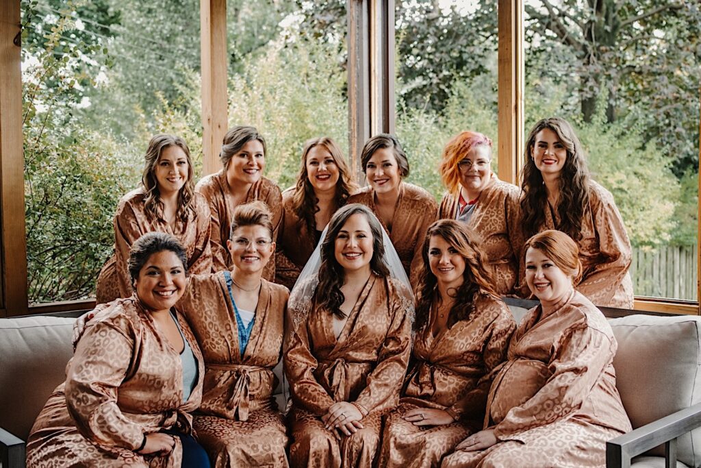A bride sits on a couch surrounded by her bridesmaids who are all wearing matching leopard print robes and smiling at the camera