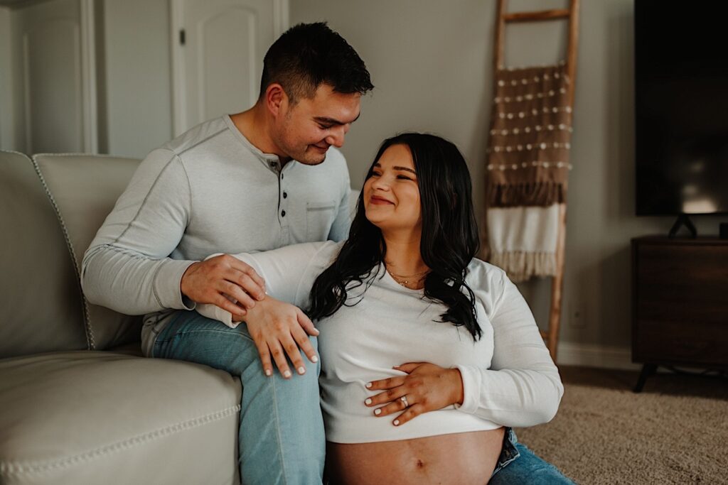 A man sits on the couch with is pregnant wife sitting in between his legs as they both smile at one another.