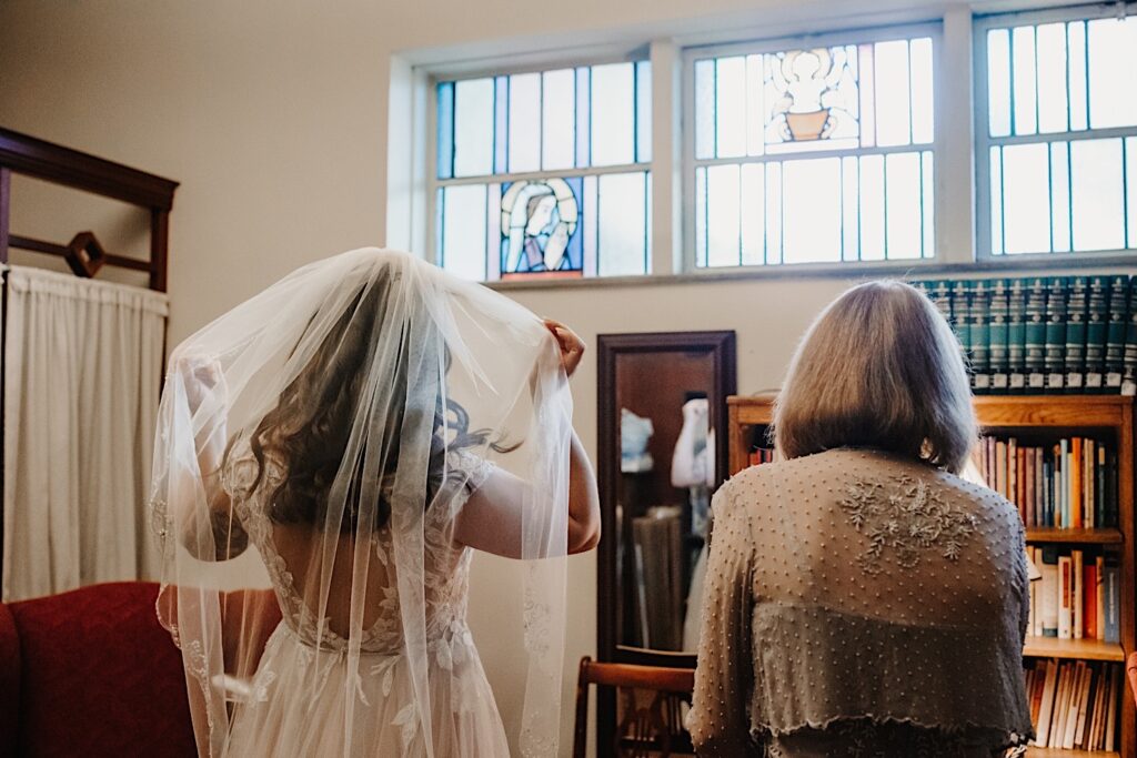 A bride and her mother stand side by side in a room of a church facing away from the camera. The bride is fluffing her veil as she gets ready for her wedding ceremony