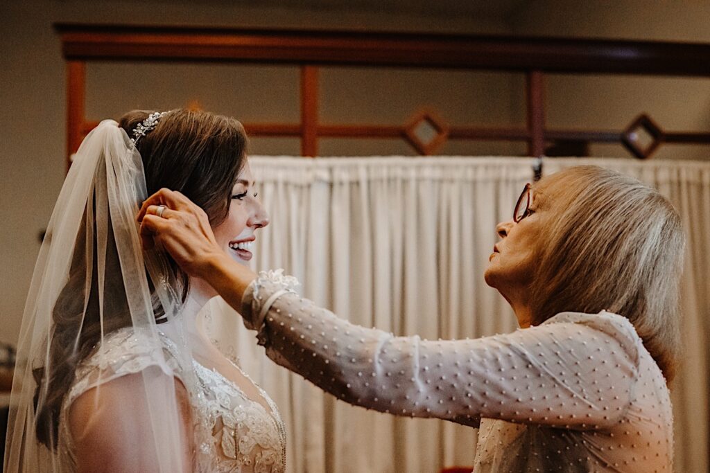 A bride smiles and laughs as her mom looks at her and helps adjust her veil before her wedding ceremony