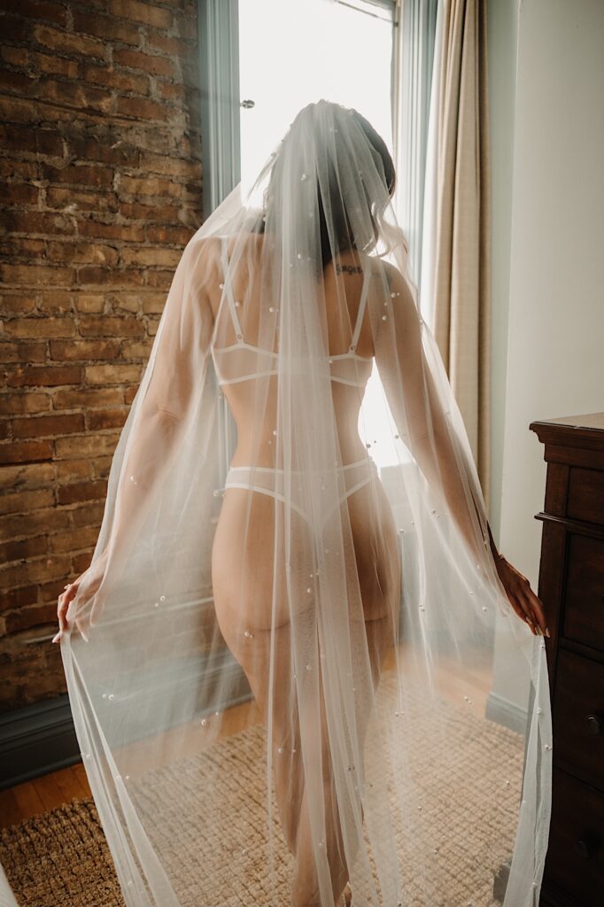 A woman wearing lingerie and a sheer cloth stands facing away from the camera during a boudoir session