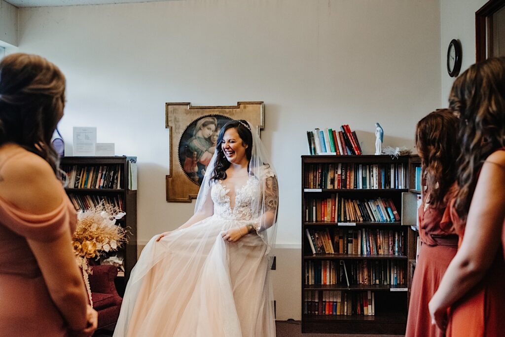 A bride smiles and plays with her dress as her bridesmaids look on in the foreground with their backs to the camera