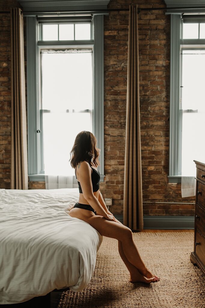 A woman in lingerie sits on the edge of a bed and looks out the window away from the camera during a boudoir session