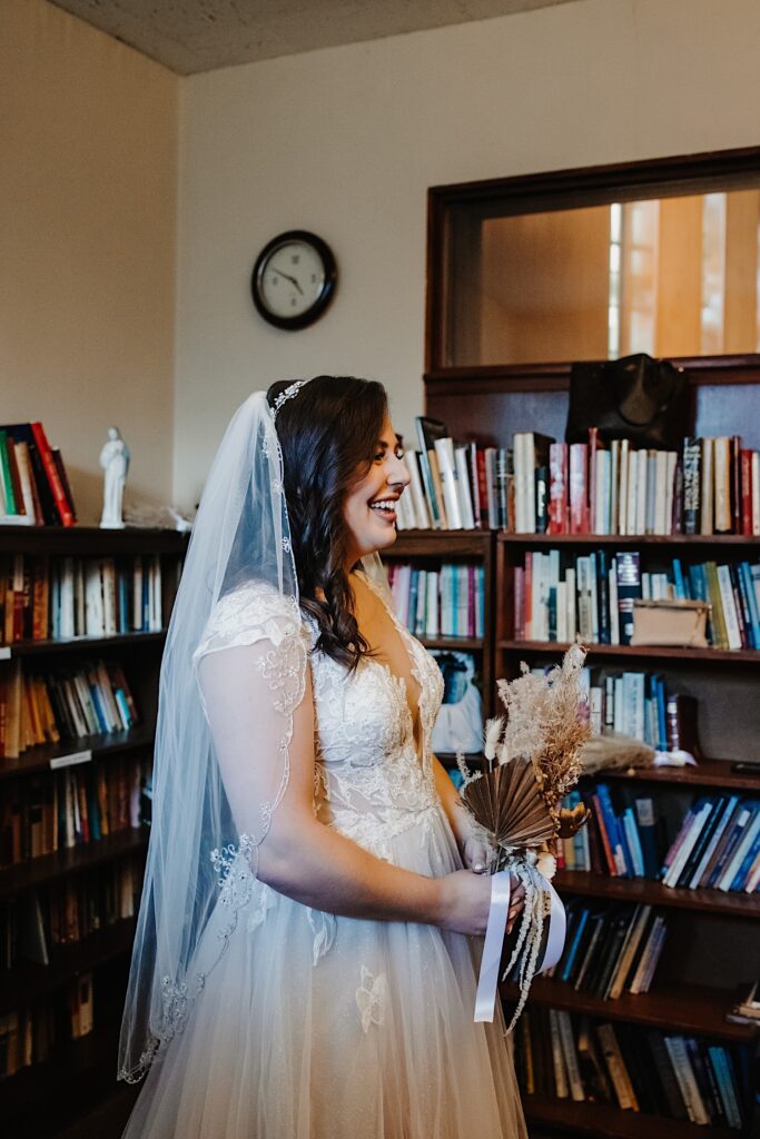 A bride in her wedding dress stands and smiles in a room full of books with her bouquet in hand