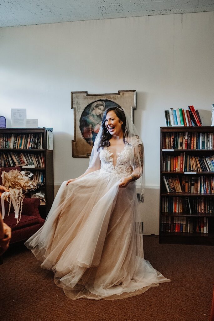 A bride smiles as she plays with her dress and stands in a room of a church before her wedding ceremony