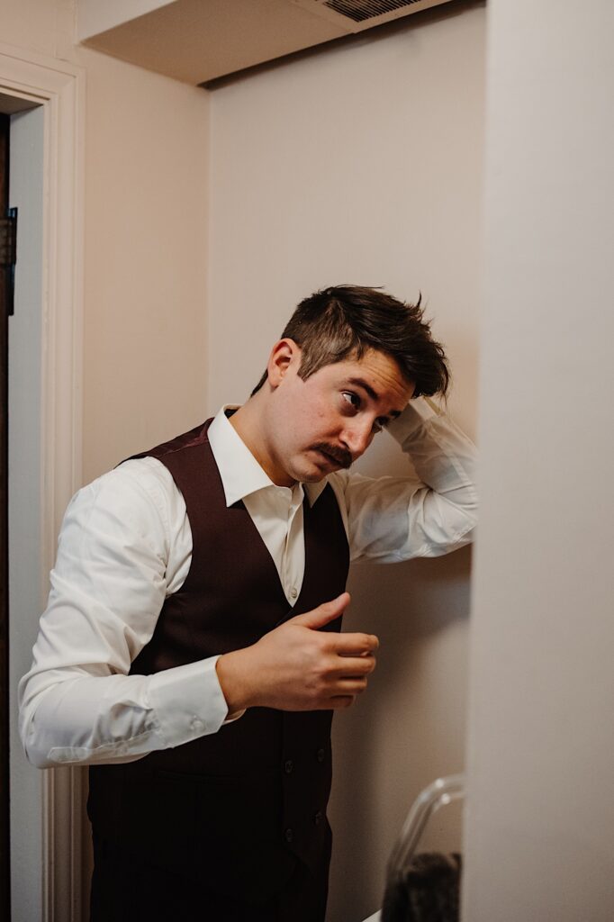A man in a dress shirt and vest adjusts his hair as he looks in the mirror