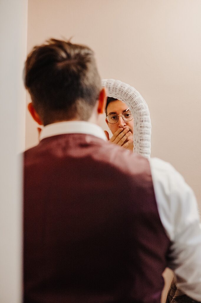A mirror in the background is in focus, in it is the face of a man touching his moustache, and in the foreground of the photo is the back of that man which is out of focus
