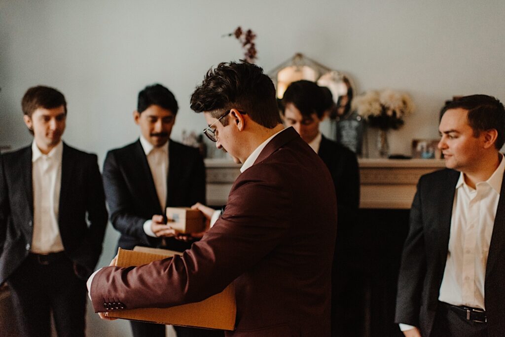 A groom stands holding a box in a room filled with his groomsmen, he is handing a smaller box to one of the groomsmen