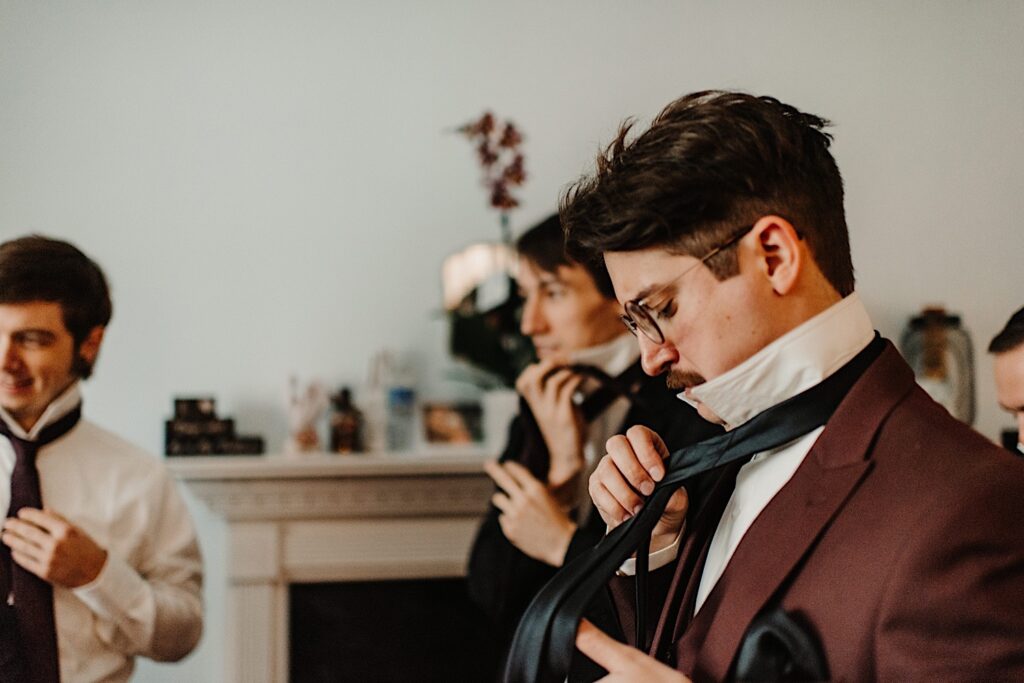 A groom stands and ties his tie as his groomsmen in the background all do the same before his wedding