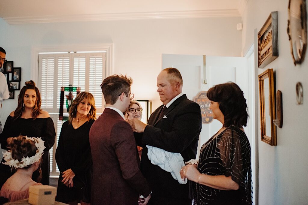 A groom stands as his father adjusts his tie in their home while the rest of his family watches