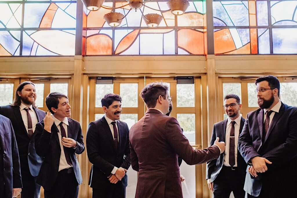 In a church under the stained glass entryway a groom talks with his groomsmen as they enter the church for his fall Chicago wedding