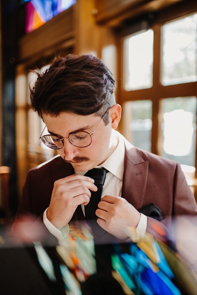 A groom stands in front of a stained glass window and adjusts his tie before his wedding ceremony