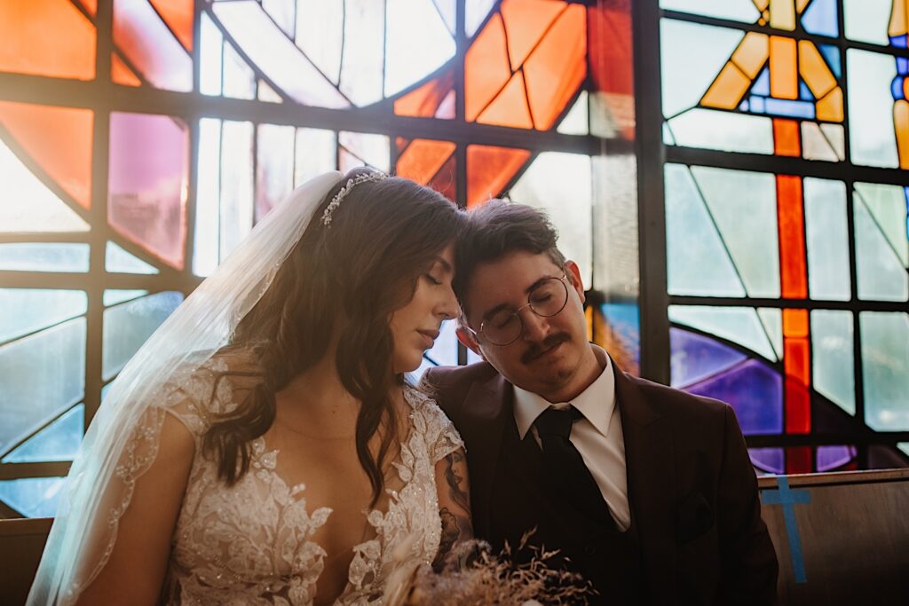 A bride and groom sit and place their foreheads together with their eyes closed during their fall wedding in Chicago, behind them is stained glass