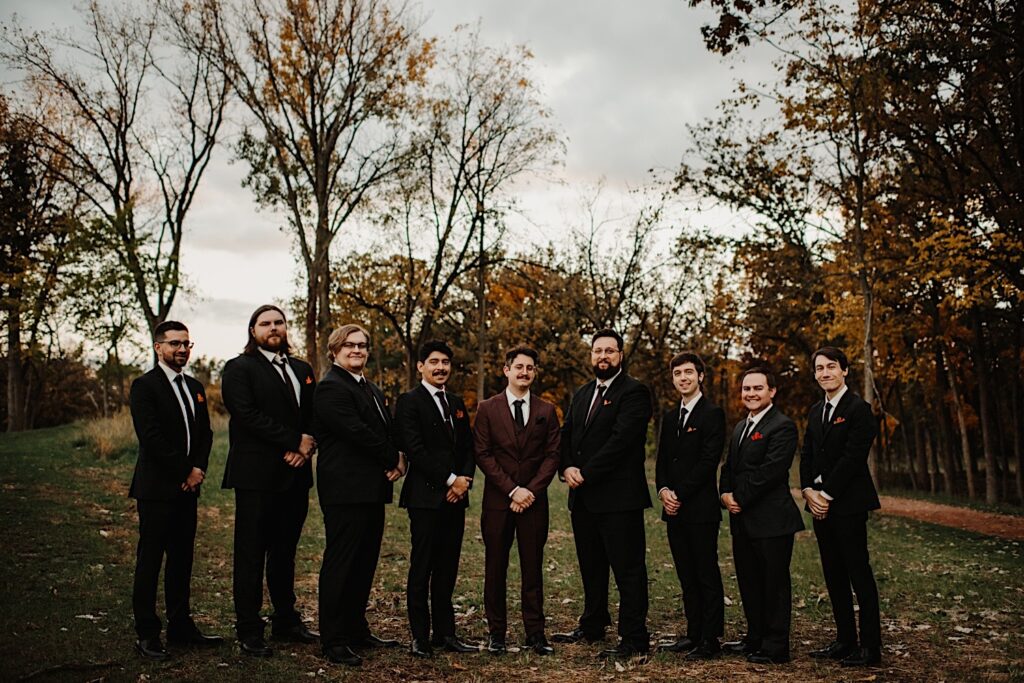 A groom and his groomsmen on either side of him stand and smile at the camera in park during a fall wedding in Chicago