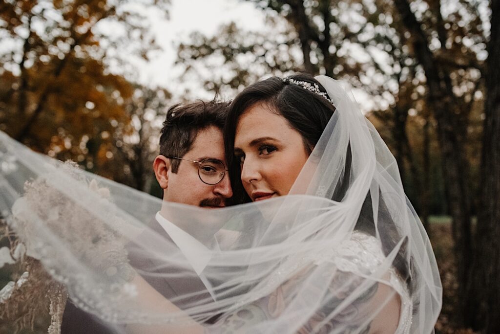A bride and groom stand in a park and embrace while looking at the camera during their fall wedding in Chicago, the brides veil flows across both of them in the foreground
