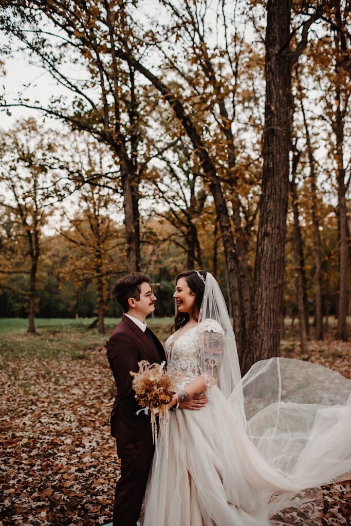 A bride and groom stand in a park filled with leaves and smile at one another on their wedding day