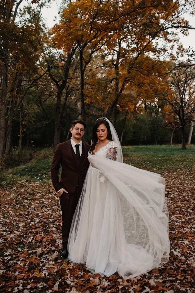 A bride and groom stand in a park filled with leaves, the groom is looking at the camera while the bride looks at and plays with her dress