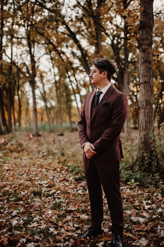A groom stands in his wedding attire looking off to the left in a park of Chicago filled with fall leaves