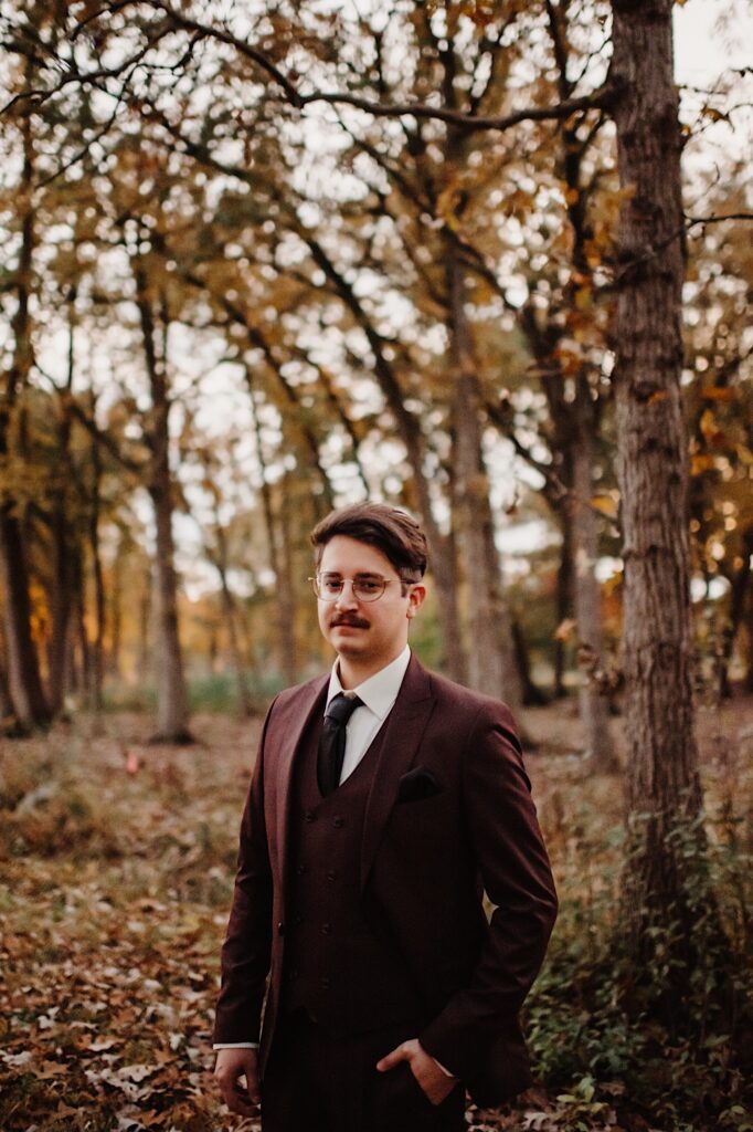 A groom in a park in the fall stands and looks at the camera while one of his hands is in his pocket