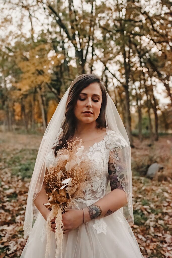 A bride stands in a park during the fall while holding her bouquet and looking down at the ground