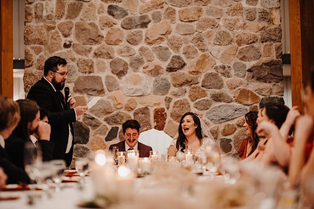 A bride and groom laugh as a groomsmen to their left gives a speech during their fall wedding reception in Chicago