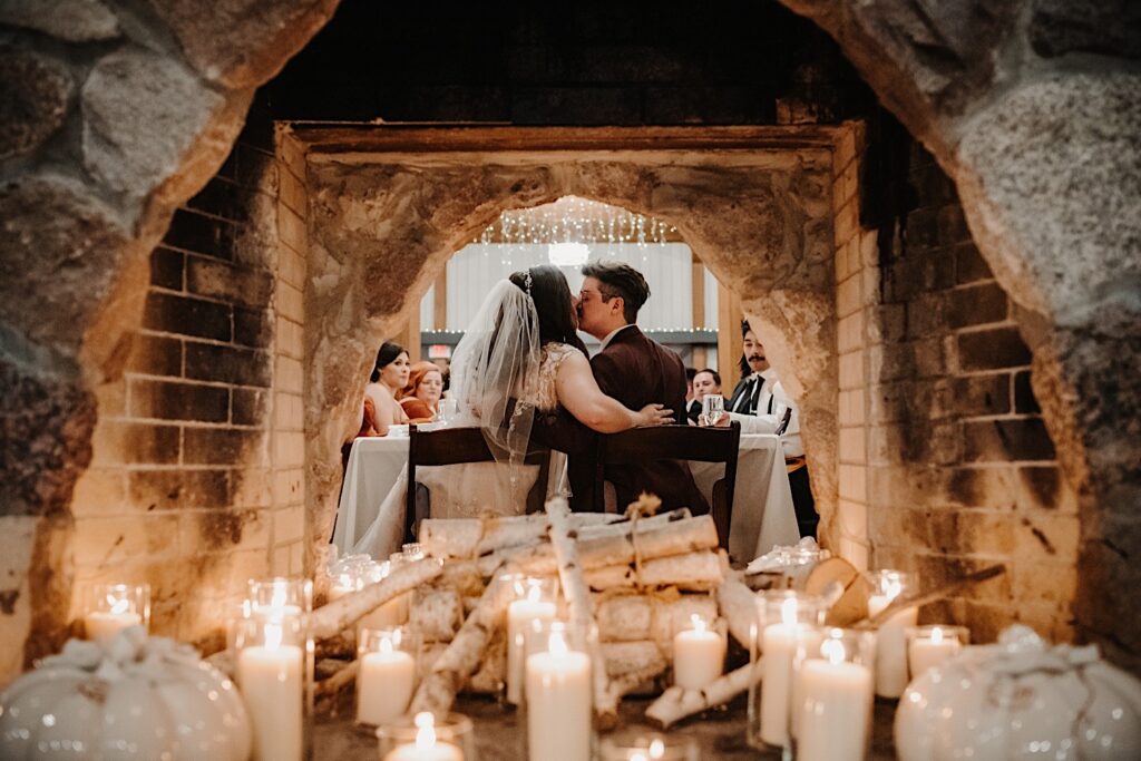 Photo of the inside of an old fireplace at Oakbrook Bath and Tennis Club lilt by candles, through the fireplace you can see the bride and groom seated and kissing during their fall wedding reception in Chicago