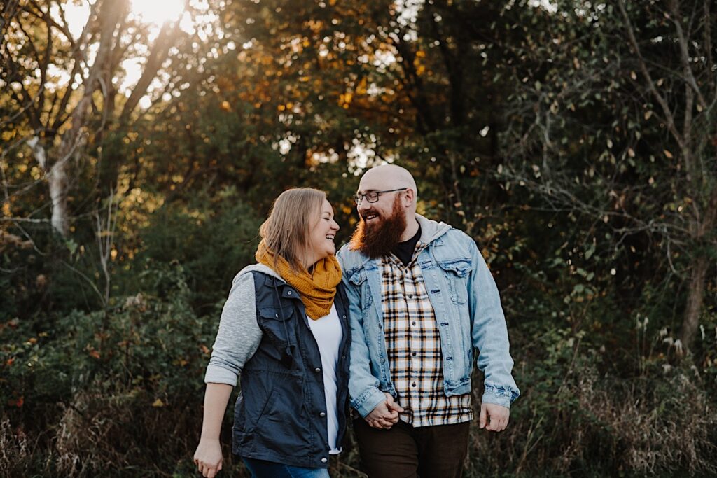 A couple laugh and smile at one another while walking in a forest in the fall, photographed by a Chicago engagement photographer