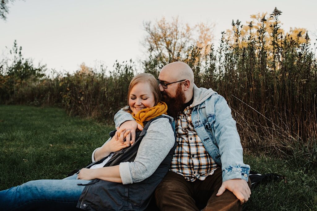 A couple sit on the grass in a field, the man whispers in the woman's ear while she laughs during their engagement session with a Chicago photographer