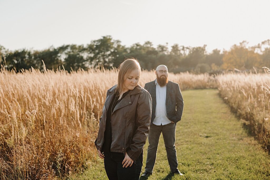 A woman stands in a field and looks down while a man stands behind her looking at her during their engagement session