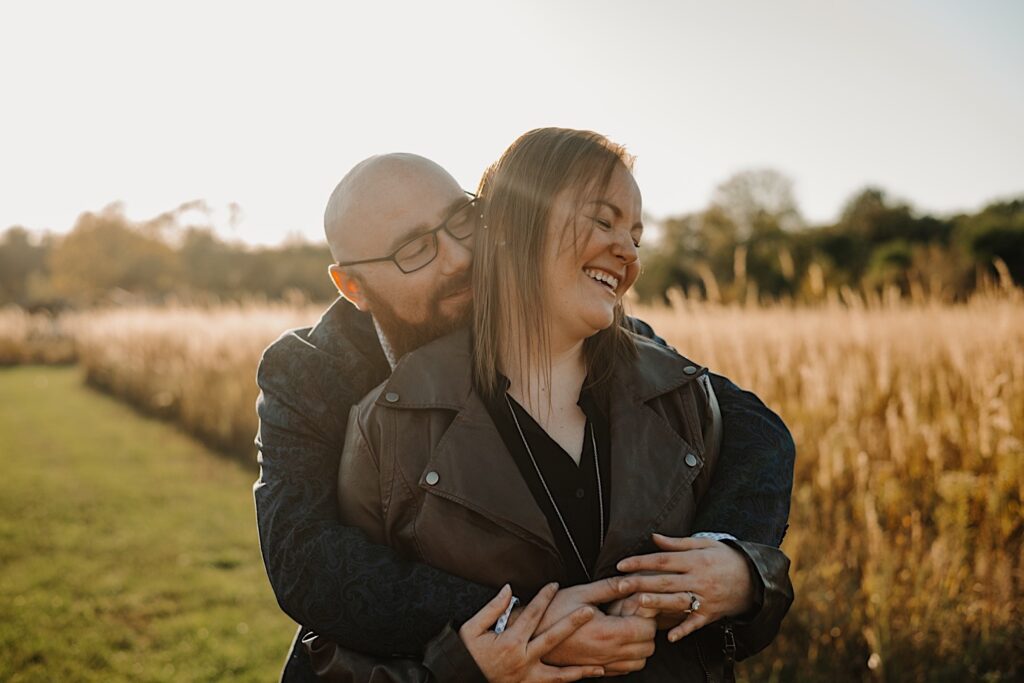 A man hugs a woman from behind as she smiles and laughs, they're in a field as the sun sets behind them, taken by a Chicago engagement photographer