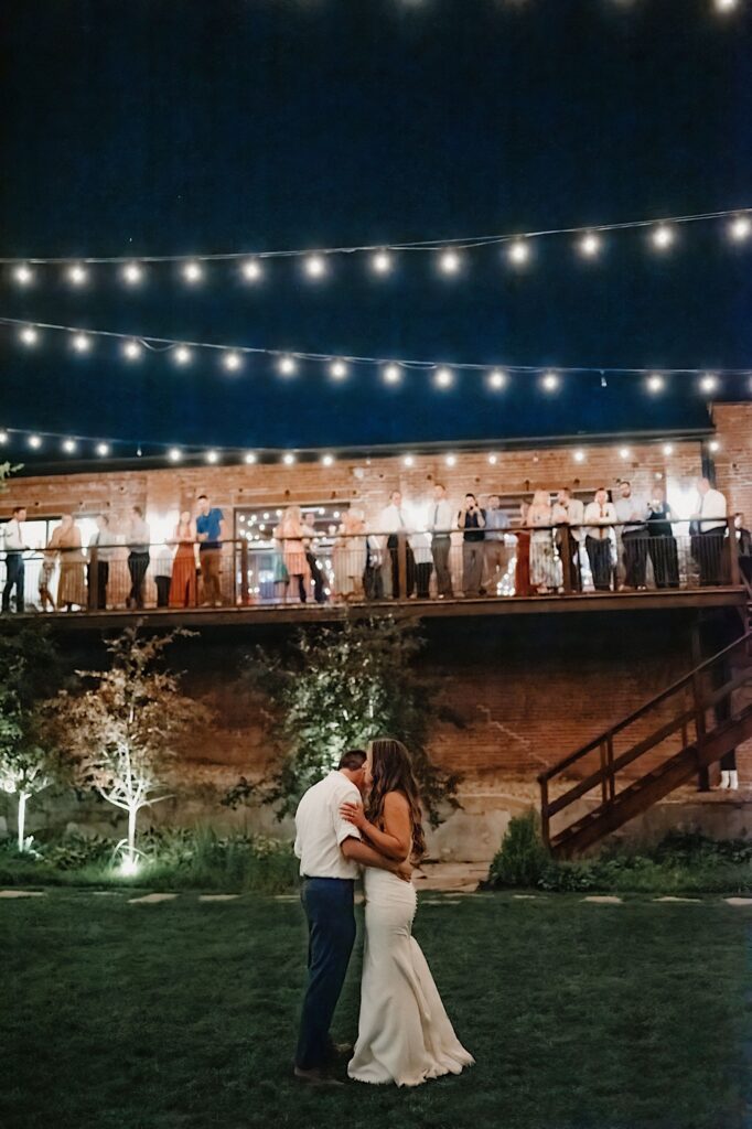 A bride and groom dance on a lawn underneath string lights while their guests stand on a balcony above them watching