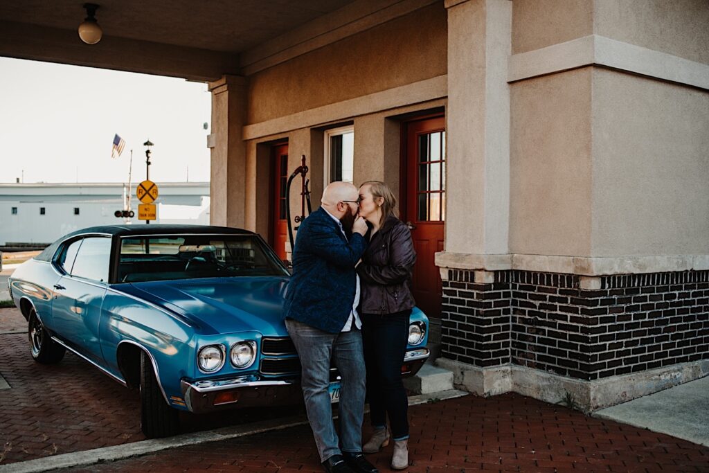 A couple kiss while standing in front of a classic Chevy SS at a retro gas station, taken by a Chicago engagement photographer