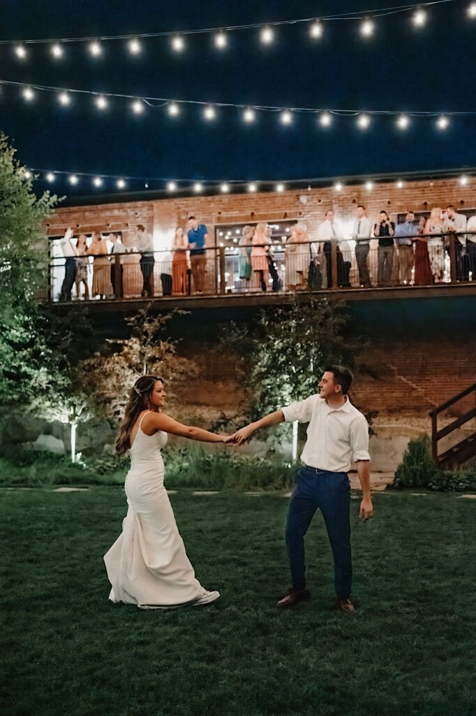 A bride and groom dance on a lawn underneath string lights while their guests stand on a balcony above them watching