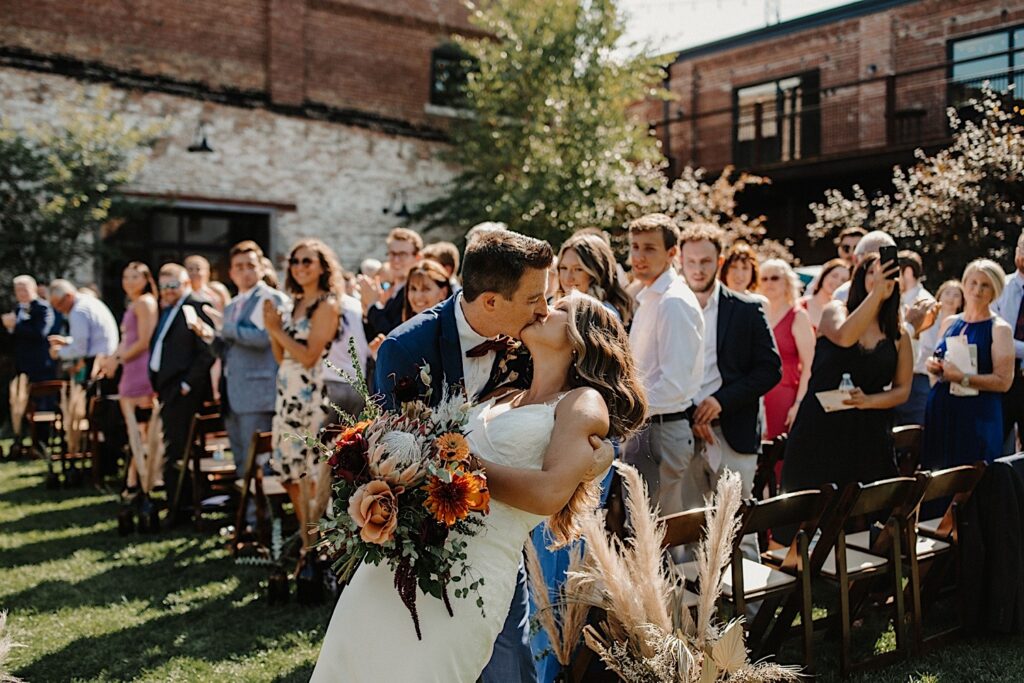 A bride and groom kiss while walking down the aisle as their guests watch and cheer for them after their outdoor wedding ceremony at The Cannery in Eureka Illinois