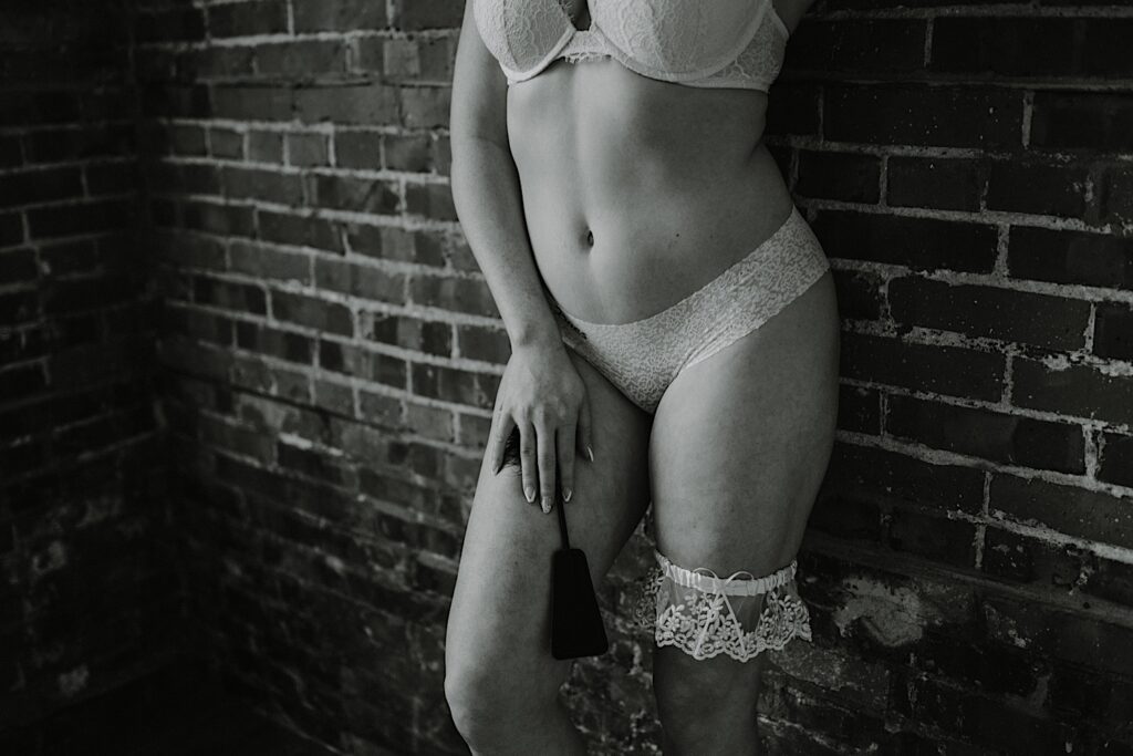 Black and white photo of a woman in lingerie standing against a brick wall during a boudoir photography session