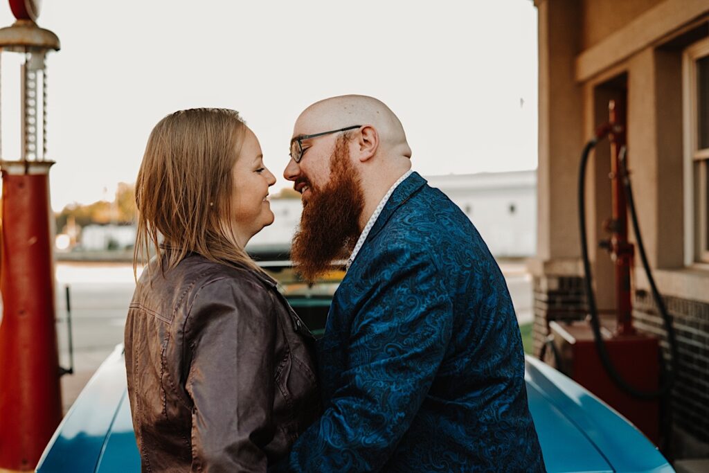 A couple stand in front of a car at a vintage gas station and smile as they lean in for a kiss