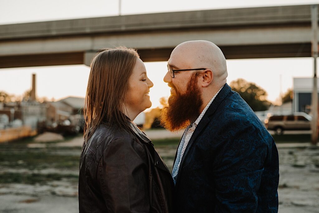 A couple smile as they lean in for a kiss while the sun sets underneath a bridge behind them