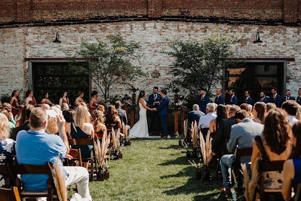 A wedding ceremony takes place in the outdoor space of the venue The Cannery in Eureka Illinois, the couple are holding hands while the officiant speaks and their guests are seated on either side of them