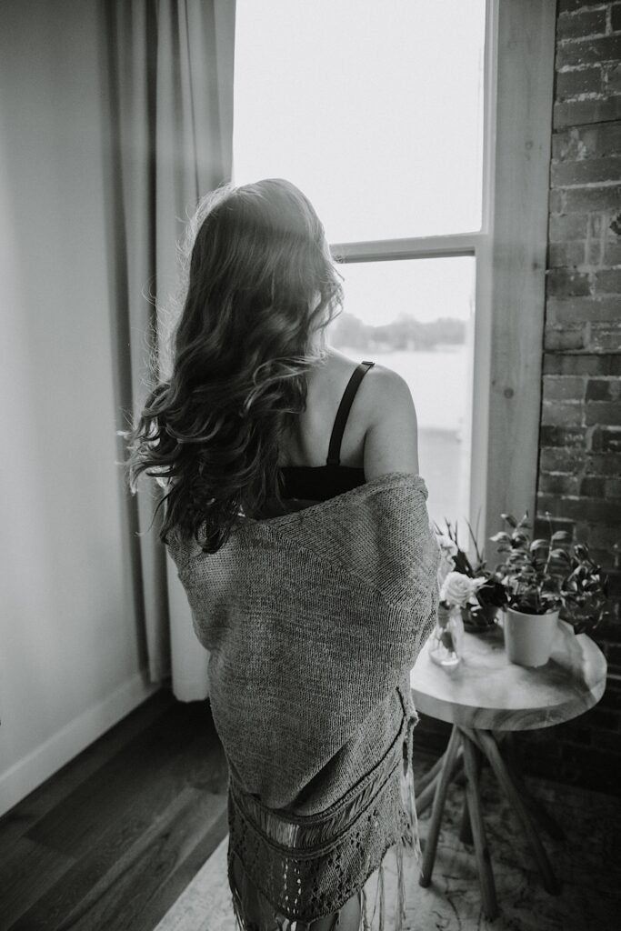 Black and white photo of a woman in lingerie facing away from the camera towards a window with a shawl draped over her back