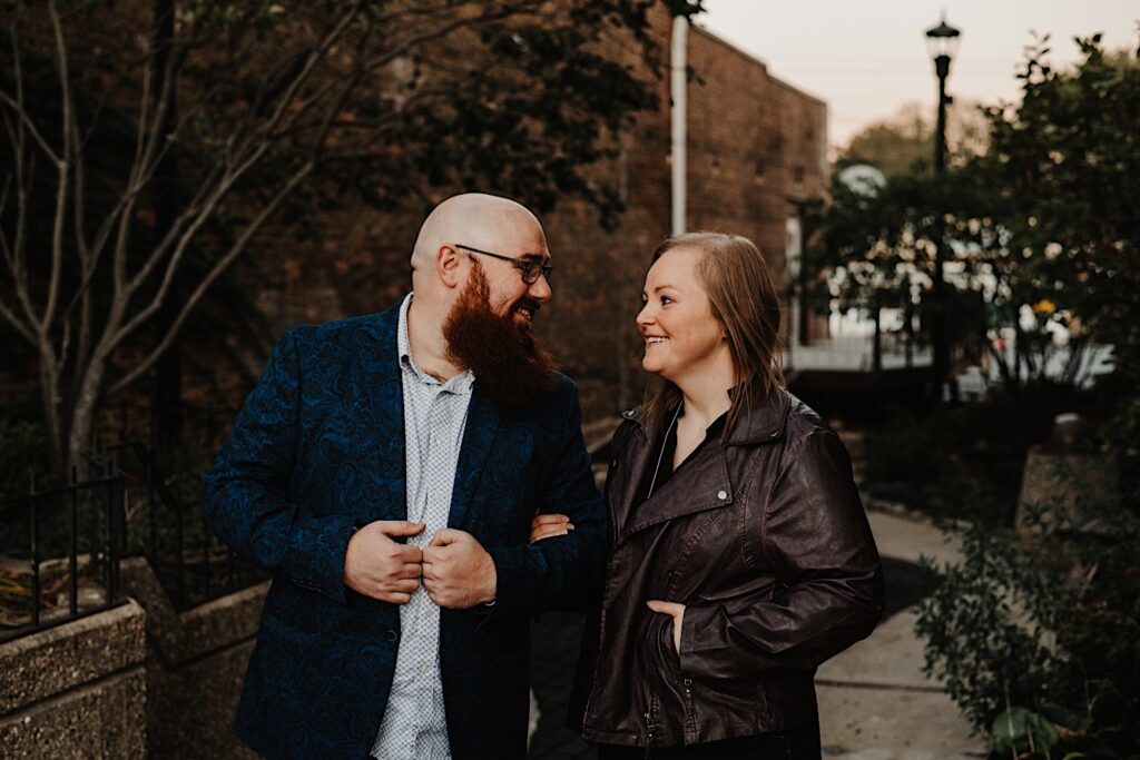 A couple walk through a park between some downtown buildings while smiling at one another, photographed by a Chicago engagement photographer