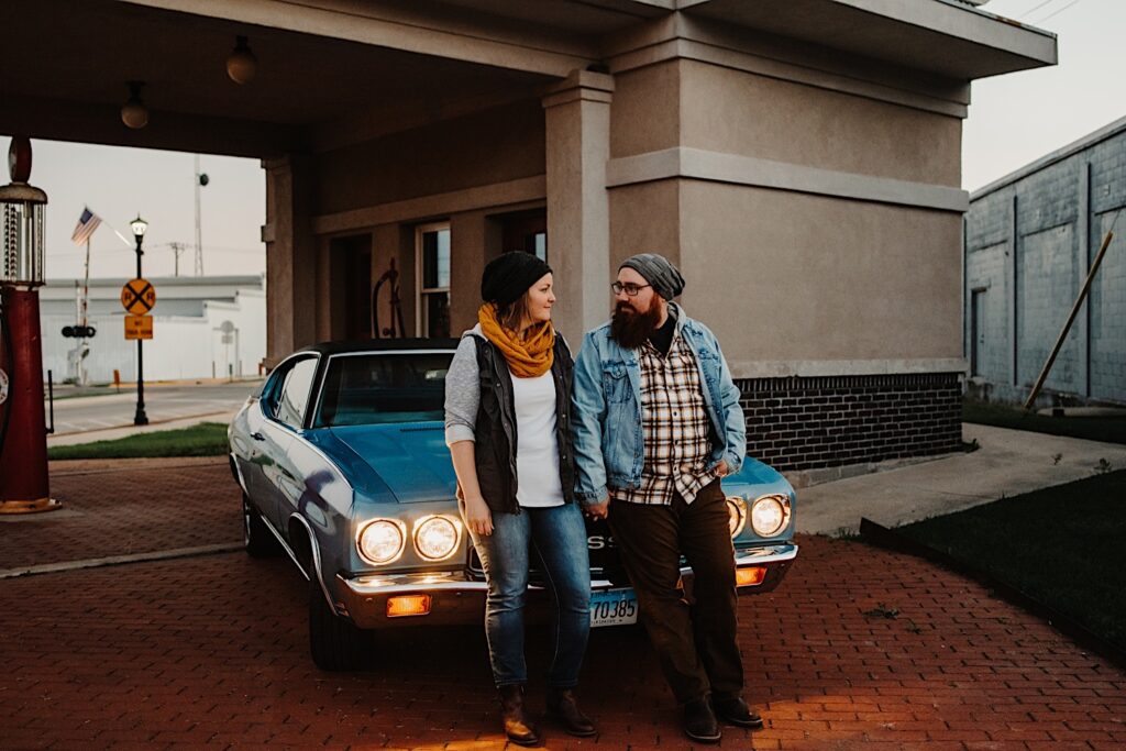 A couple in their fall clothes hold hands and lean against their classic Chevy SS with it's headlights on at a vintage gas station, photographed by a Chicago engagement photographer