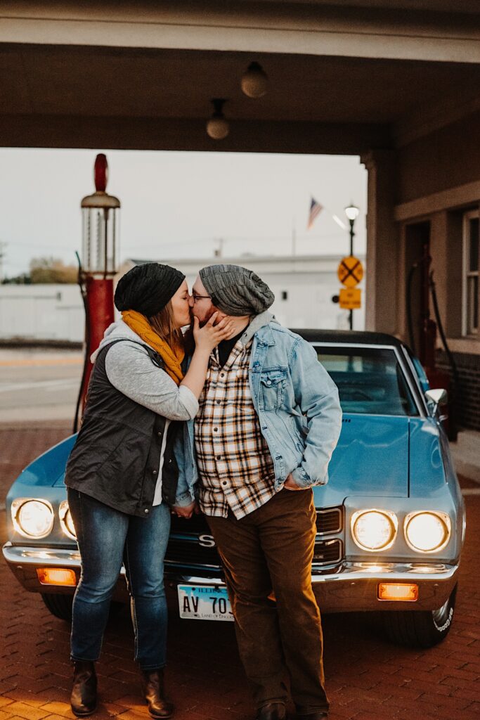 A couple in their fall clothes kiss in front of their classic Chevy SS with it's headlights on at a vintage gas station