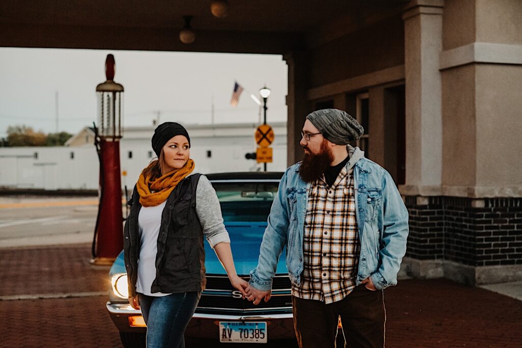 A couple in their fall clothes hold hands and stand in front of their classic Chevy SS with it's headlights on at a vintage gas station, photographed by a Chicago engagement photographer