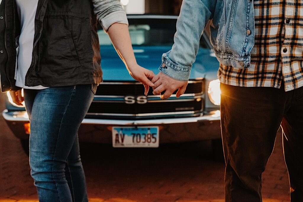Close up photo of a couple in their fall clothes holding hands and stand in front of their classic Chevy SS with it's headlights on at a vintage gas station, photographed by a Chicago engagement photographer
