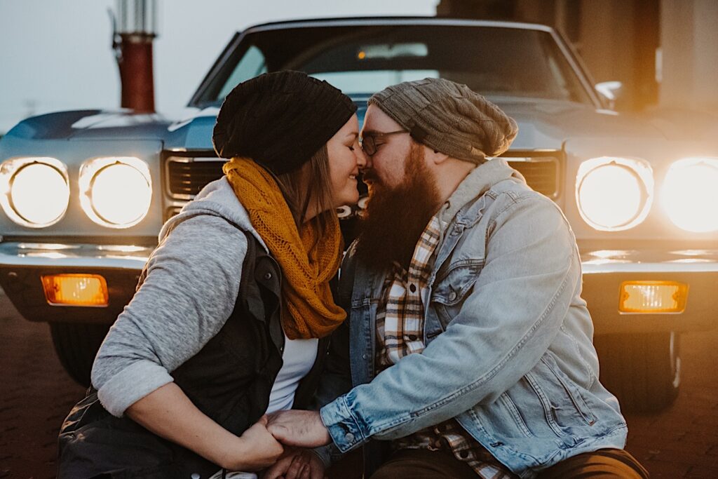 A couple in their fall clothes sit in front of their classic Chevy SS with its headlights on and lean in for a kiss while at a vintage gas station, photographed by a Chicago engagement photographer